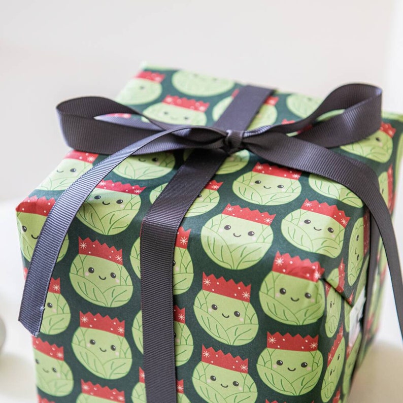 Pode incluir: Uma caixa de presente verde com uma fita preta. A caixa &eacute; embrulhada em papel com um padr&atilde;o de br&oacute;colis de desenho animado usando chap&eacute;us de Papai Noel.