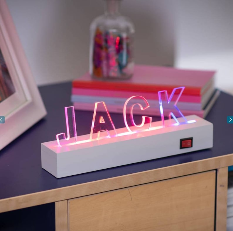 May include: A white light-up sign with the name "JACK" spelled out in clear acrylic letters. The letters are illuminated with pink and blue LED lights. The sign is on a white wooden base with an on/off switch.