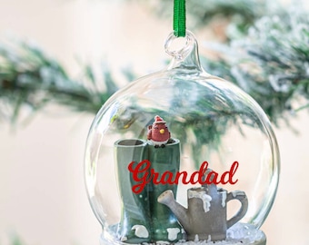 May include: A glass Christmas ornament with a red cardinal, green boots, and a gray watering can inside. The ornament has the word "Grandad" written on it in red.