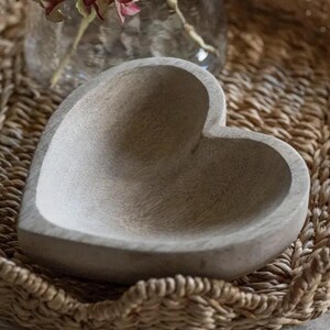 May include: A heart-shaped bowl carved from light-colored wood with a visible grain pattern. The bowl features a smooth, concave interior and a thick, rounded rim, presenting a natural, unpainted finish with a matte texture throughout the surface.