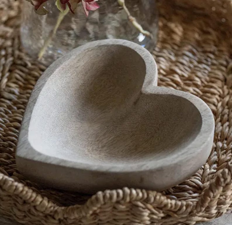 May include: A heart-shaped bowl carved from light-colored wood with a visible grain pattern. The bowl features a smooth, concave interior and a thick, rounded rim, presenting a natural, unpainted finish with a matte texture throughout the surface.