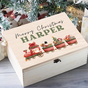 Könnte beinhalten: Eine hölzerne Weihnachts-Erinnerungskiste mit dem personalisierten Namen "HARPER" und dem Text "Frohe Weihnachten". Der Deckel zeigt eine farbenfrohe Illustration eines Zuges mit dem Weihnachtsmann, einem Schneemann und anderen festlichen Figuren.