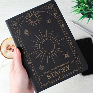 May include: Black journal with a gold design featuring a sun, moon, and stars. The text "Follow Your Dreams" and the name "Stacey" are also included in the design.