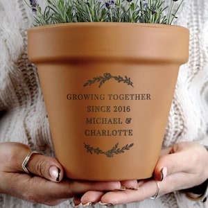 May include: A terracotta-colored flower pot with the text "GROWING TOGETHER SINCE 2016 MICHAEL & CHARLOTTE" and floral accents. The pot is filled with lavender plants. The pot is being held by a person.