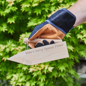 Könnte beinhalten: Ein hölzerner Beerensammler mit einem Metallgriff. Der Sammler ist mit dem Text "Mike's Berry Special Picker" graviert.