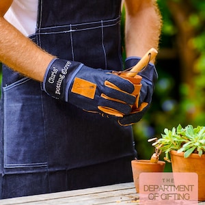 以下が含まれることがあります： タン革のアクセントと「Chris' potting glove」の文字が入った濃い青色の植木鉢用手袋。濃い青色のエプロンを着た人が手袋を持っています。小さなテラコッタの鉢と植物が背景にあります。
