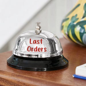 Puede incluir: Una campana cromada brillante con una base negra. Las palabras "Last Orders" est&aacute;n impresas en rojo en la superficie de la campana. La campana est&aacute; sobre una superficie de madera, con un jarr&oacute;n decorativo en el fondo.