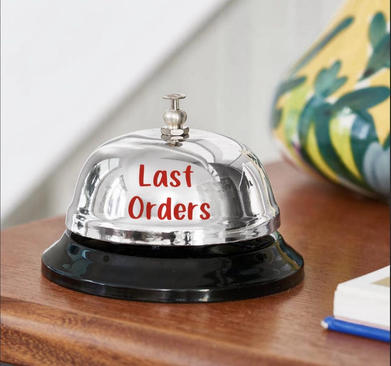 Puede incluir: Una campana cromada brillante con una base negra. Las palabras "Last Orders" est&aacute;n impresas en rojo en la superficie de la campana. La campana est&aacute; sobre una superficie de madera, con un jarr&oacute;n decorativo en el fondo.