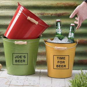 Op de afbeelding: Drie metalen emmers in rood, groen en geel. Op de groene emmer staat "JOE'S BEER". De gele emmer heeft "TIME FOR BEER" erop gedrukt en bevat twee groene bierflessen in ijs. Elke emmer heeft een houten handvat.