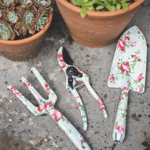 K&ouml;nnte beinhalten: Ein Gartenger&auml;teset mit einer Kelle, einer Handgabel und einer Gartenschere. Jedes Werkzeug hat einen hellblauen Hintergrund mit einem Blumenmuster aus roten und rosa Rosen. Die Werkzeuge liegen auf einer grauen Oberfl&auml;che.