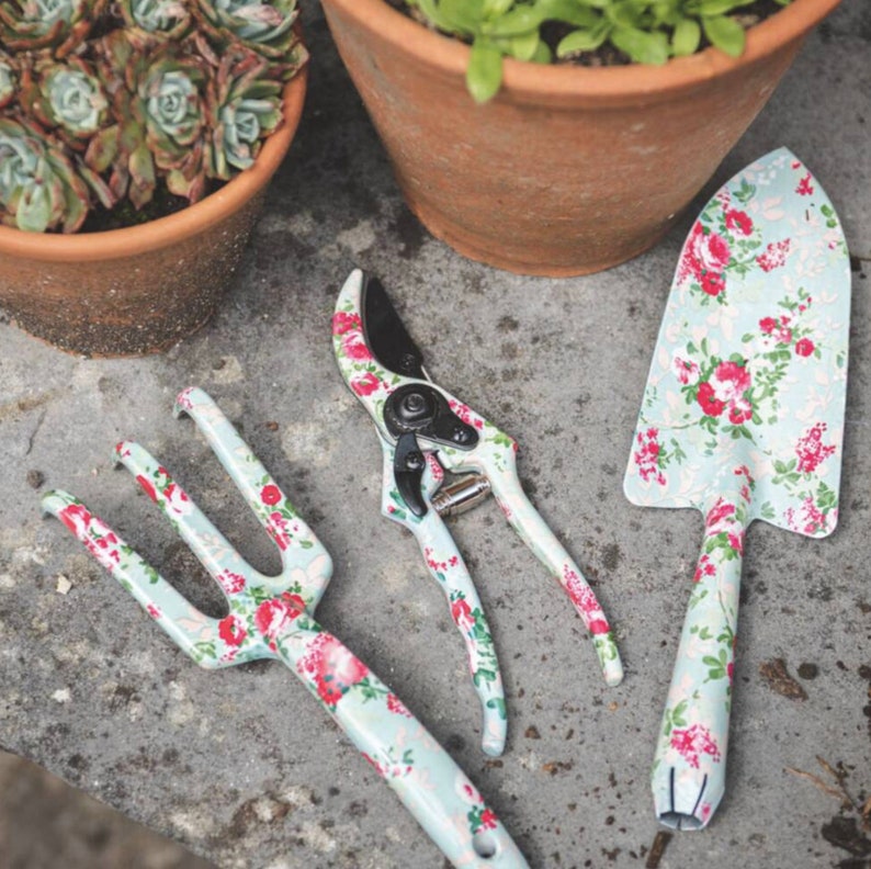 K&ouml;nnte beinhalten: Ein Gartenger&auml;teset mit einer Kelle, einer Handgabel und einer Gartenschere. Jedes Werkzeug hat einen hellblauen Hintergrund mit einem Blumenmuster aus roten und rosa Rosen. Die Werkzeuge liegen auf einer grauen Oberfl&auml;che.