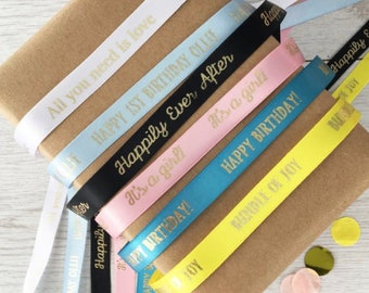 May include: Assortment of colorful satin ribbon with gold lettering. The ribbons have various messages including "Happy Birthday", "Happily Ever After", "It's a Girl", and "All You Need is Love".