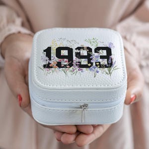 Op de afbeelding: Een witte, vierkante sieradendoos met het jaartal "1993" in grote zwarte cijfers. De doos is versierd met een bloemenontwerp in verschillende kleuren en heeft een zilveren ritssluiting. De doos wordt in open handen gehouden.