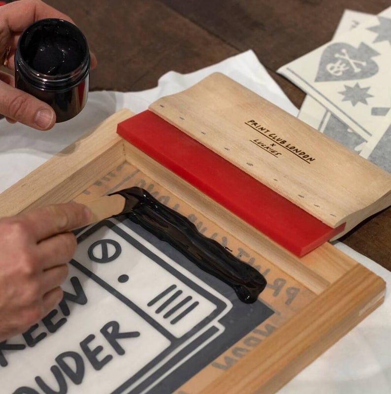 K&ouml;nnte beinhalten: Ein Holzrahmen f&uuml;r den Siebdruck mit einem roten Rakel und schwarzer Tinte, die auf ein graues T-Shirt mit dem aufgedruckten Text "Screen Sluder" aufgetragen wird. Der Rahmen tr&auml;gt ein Etikett mit der Aufschrift "Paint Club London x Luckies".