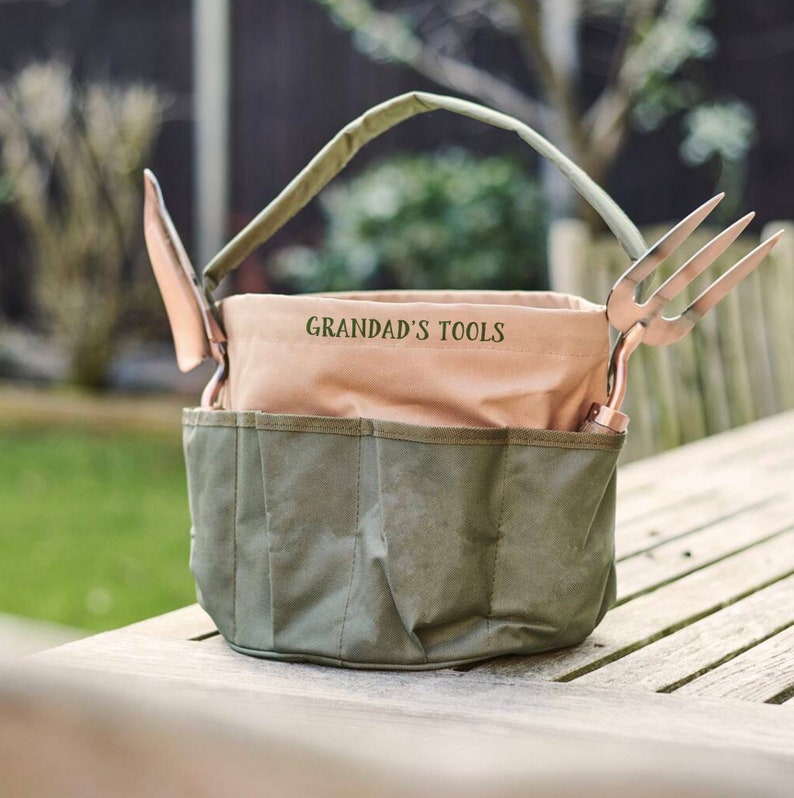 K&ouml;nnte beinhalten: Eine gr&uuml;ne und beige Canvas-Gartentasche mit Griff und Taschen. Die Tasche enth&auml;lt eine Kelle und eine dreizackige Gabel. Der Text "GRANDAD'S TOOLS" ist auf der Vorderseite der Tasche aufgedruckt.