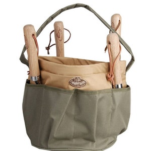 K&ouml;nnte beinhalten: Eine Gartentasche aus olivgr&uuml;nem und beigefarbenem Canvas mit Griff und mehreren Taschen. Sie enth&auml;lt vier Gartenger&auml;te mit Holzgriffen und Lederschlaufen. Die Tasche hat ein Etikett mit der Aufschrift "Orchard & Garden".