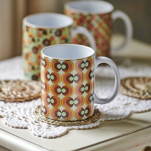 May include: Three ceramic mugs with retro geometric patterns in shades of brown, orange, and green. The mugs are on a white crocheted doily with woven coasters.
