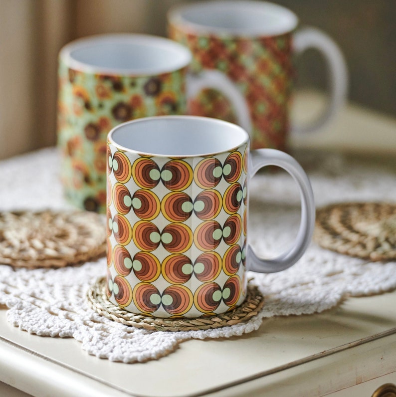 May include: Three ceramic mugs with retro geometric patterns in shades of brown, orange, and green. The mugs are on a white crocheted doily with woven coasters.