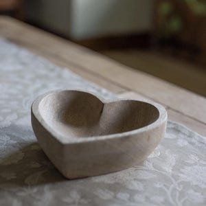 May include: A heart-shaped bowl carved from light-colored wood with a visible grain pattern. The bowl features a smooth, rounded interior basin and a thick, sturdy rim, showcasing the natural texture and neutral beige tones of the material.