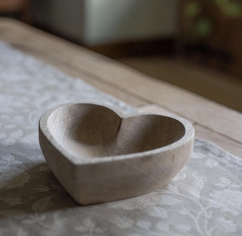 May include: A heart-shaped bowl carved from light-colored wood with a visible grain pattern. The bowl features a smooth, rounded interior basin and a thick, sturdy rim, showcasing the natural texture and neutral beige tones of the material.