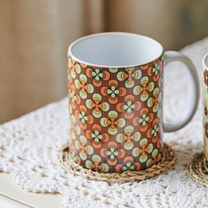 May include: A ceramic mug with a retro floral pattern in shades of brown, orange, green, and yellow. The mug has a white interior and a handle. It is sitting on a woven coaster.