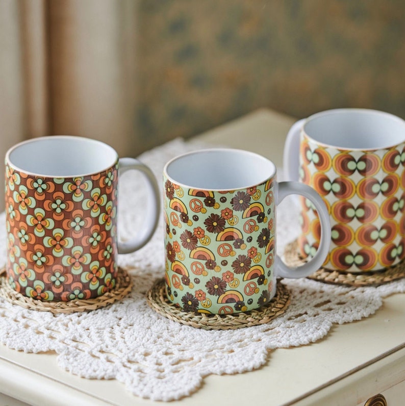 May include: Three ceramic mugs with retro patterns. The mugs are on a white crocheted doily. The mugs have different patterns, including flowers, peace signs, and geometric shapes. The mugs are in shades of brown, orange, green, and white.