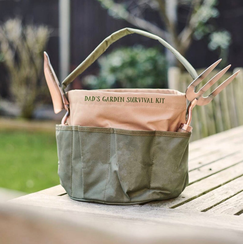 K&ouml;nnte beinhalten: Eine gr&uuml;ne und beige Canvas-Gartentasche mit Griff, die eine Kelle und eine Gartenforke enth&auml;lt. Die Tasche hat mehrere Taschen und den Text "DAD'S GARDEN SURVIVAL KIT". Die Werkzeuge haben kupferfarbene Metallk&ouml;pfe.