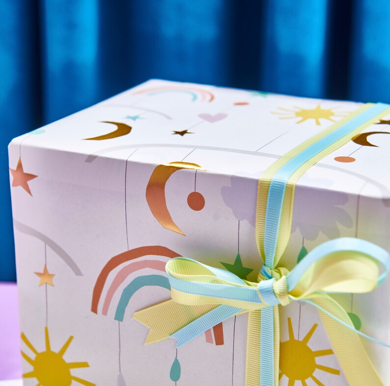 K&ouml;nnte beinhalten: Eine wei&szlig;e Geschenkbox mit einem bunten Design aus Regenb&ouml;gen, Sternen, Monden und Sonnen. Die Box ist mit einem blauen und gelben Band gebunden.