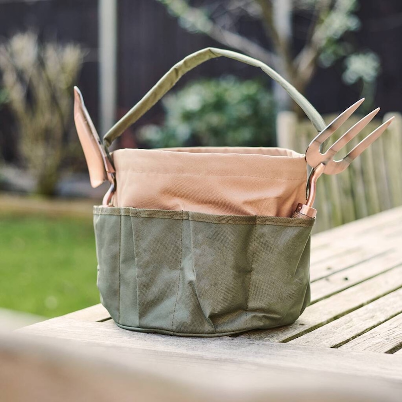 K&ouml;nnte beinhalten: Eine gr&uuml;ne und beige Canvas-Gartenger&auml;tetasche mit Griff und Taschen. Die Tasche enth&auml;lt eine kupferfarbene Kelle und eine Gartenforke. Die Tasche steht auf einem Holztisch.