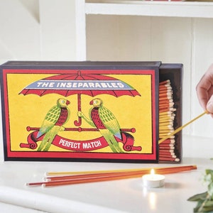 May include: A red, white, and blue matchbox with a yellow background. The matchbox features two green parrots under a red umbrella with the text "The Inseparables. Perfect Match".