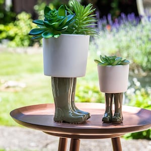 Puede incluir: Dos macetas de cerámica con forma de botas de lluvia verdes, cada una con una planta suculenta. Las macetas están sobre una mesa de color cobre.