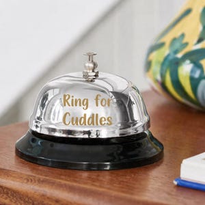 Puede incluir: Una campana plateada brillante con una base negra. La campana tiene la inscripci&oacute;n dorada "Ring for Cuddles". Est&aacute; colocada sobre una superficie de madera.
