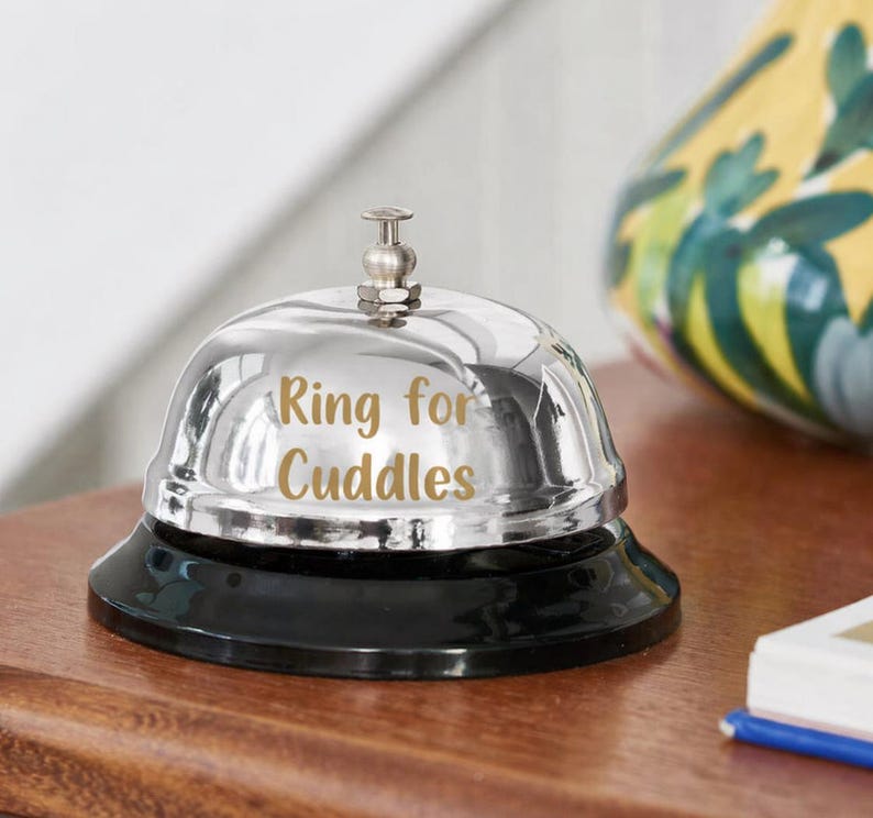 Puede incluir: Una campana plateada brillante con una base negra. La campana tiene la inscripci&oacute;n dorada "Ring for Cuddles". Est&aacute; colocada sobre una superficie de madera.