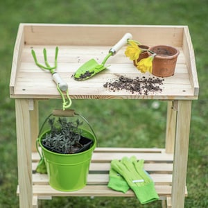Può includere: Un banco da giardinaggio in legno con attrezzi e forniture per il giardinaggio. Il banco contiene una cazzuola verde, una forca a mano verde, un secchio verde con una pianta, un paio di guanti verdi e due piccoli vasi di terracotta con fiori gialli. Il terreno è sparso sul banco.