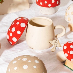 May include: A beige ceramic mug with a curved handle and a mushroom-shaped tag that reads "Oliva's Tea." The mug is surrounded by mushroom-shaped decor, including red and white spotted mushroom lids and a mushroom-shaped base.