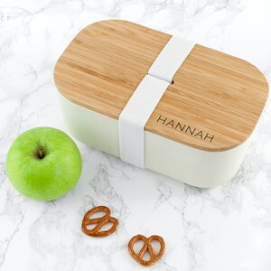 Puede incluir: Una fiambrera de bambú natural y blanco con una correa elástica blanca y el nombre "HANNAH" grabado en la tapa. La fiambrera está sobre una superficie de mármol blanco con una manzana verde y dos aperitivos de pretzel.