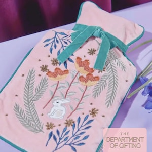 May include: A pink hot water bottle with embroidered floral and bunny design. The bottle has a teal green trim and a teal green ribbon tied at the top. The text "THE DEPARTMENT OF GIFTING" is in the bottom right corner.