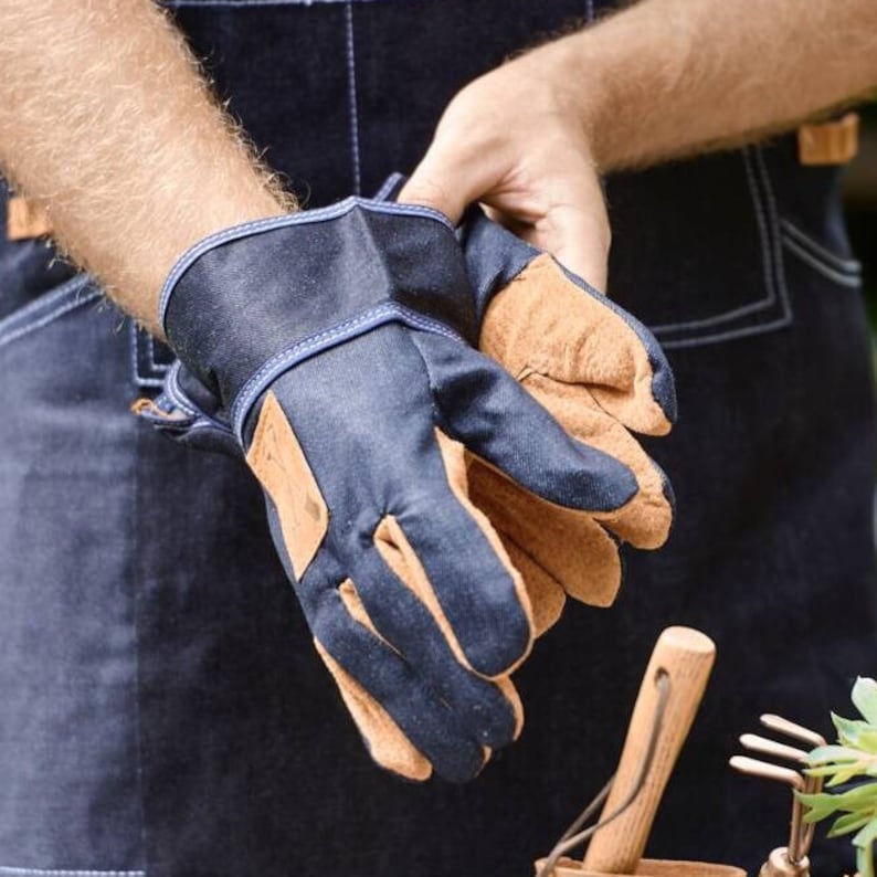 May include: A pair of blue denim gardening gloves with brown leather palms. The gloves are worn by a person wearing blue denim overalls.