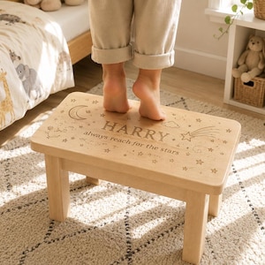 May include: A light-colored wooden step stool featuring an engraved design of a crescent moon, stars, a cloud, and a shooting star. The text "HARRY" and "always reach for the stars" is centered on the top surface of the rectangular wooden stool.
