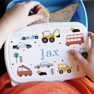 Könnte beinhalten: Weiße Lunchbox mit einem bunten Design aus verschiedenen Fahrzeugen, darunter ein Polizeiauto, ein Feuerwehrauto, ein Bus und ein Bagger. Die Lunchbox trägt den Namen "Jax" auf der Vorderseite.
