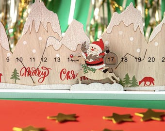 Può includere: Calendario dell'Avvento natalizio in legno a forma di catena montuosa, con numeri e le parole "Merry Christmas" in rosso. È presente Babbo Natale su una renna. Decorazioni a forma di stella dorata sparse su una superficie rossa.