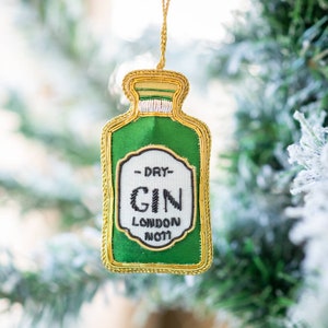 May include: A green and gold embroidered ornament shaped like a gin bottle. The ornament has the words "DRY GIN LONDON N011" on the front. The ornament is hanging from a gold string.