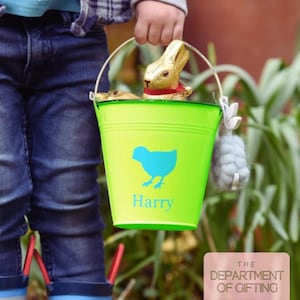 Peut inclure: Un seau de Pâques vert vif avec un motif de poussin bleu et le nom "Harry" imprimé dessus. Le seau contient un lapin en chocolat enveloppé de papier doré et d'autres friandises. Un petit pompon gris est attaché à la poignée.