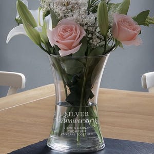 Könnte beinhalten: Eine klare Glasvase mit einer ausgestellten Öffnung und einem runden Boden. Die Vase ist mit dem Text "SILVER Anniversary Steve & Hannah Thompson 25 Years Together" graviert. Die Vase ist mit Wasser und rosa Rosen, weißen Lilien und weißen Schleierkraut gefüllt.