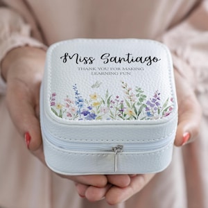 Puede incluir: Una caja de joyería cuadrada blanca con un diseño floral y el texto "Miss Santiago THANK YOU FOR MAKING LEARNING FUN". La caja tiene un cierre de cremallera y se sostiene en manos abiertas.