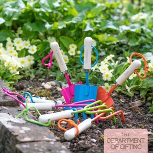 May include: A selection of brightly coloured garden tools, including trowels and hand rakes, with white handles and coloured accents. Some tools are personalised with names. The tools are arranged on soil with a backdrop of green foliage and flowers.