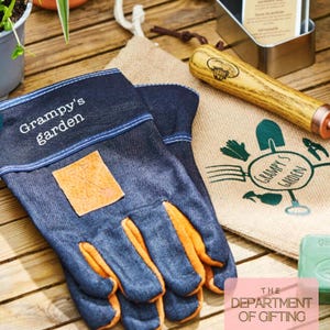 以下が含まれることがあります： オレンジ色の革の手のひらと「Grampy's garden」の文字が入ったデニムガーデニンググローブ。緑色のグラフィックと「Grampy's Garden」の文字が入ったジュートバッグが、木製の柄のガーデニングツールと金属製の容器の隣にあります。