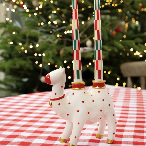 May include: A whimsical white camel-shaped candle holder with red polka dots and gold accents. It holds two tall, striped candles in red, green, and white. The candle holder sits on a red and white checkered tablecloth, with a Christmas tree in the background.