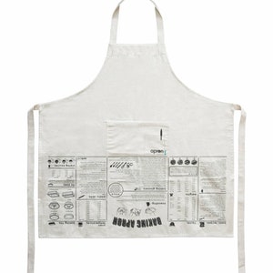 Puede incluir: Un delantal de algod&oacute;n blanco con un bolsillo grande en la parte delantera. El delantal tiene un estampado en blanco y negro de t&eacute;rminos y herramientas de reposter&iacute;a, incluyendo "Baking Apron", "temperatures", "baking times", "kneading", "pastry skills", "baking glossary", y "piping nozzles".