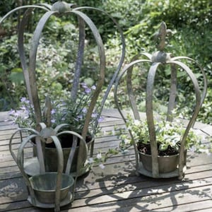 Puede incluir: Tres soportes para plantas de metal en forma de corona con un acabado desgastado. Cada soporte tiene una maceta redonda y está lleno de plantas verdes. Los soportes están sobre una superficie de madera.